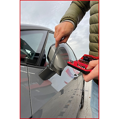 Person montiert Seitenspiegel an einem grauen Auto. In der Hand ein roter Werkzeugkasten mit Bits.